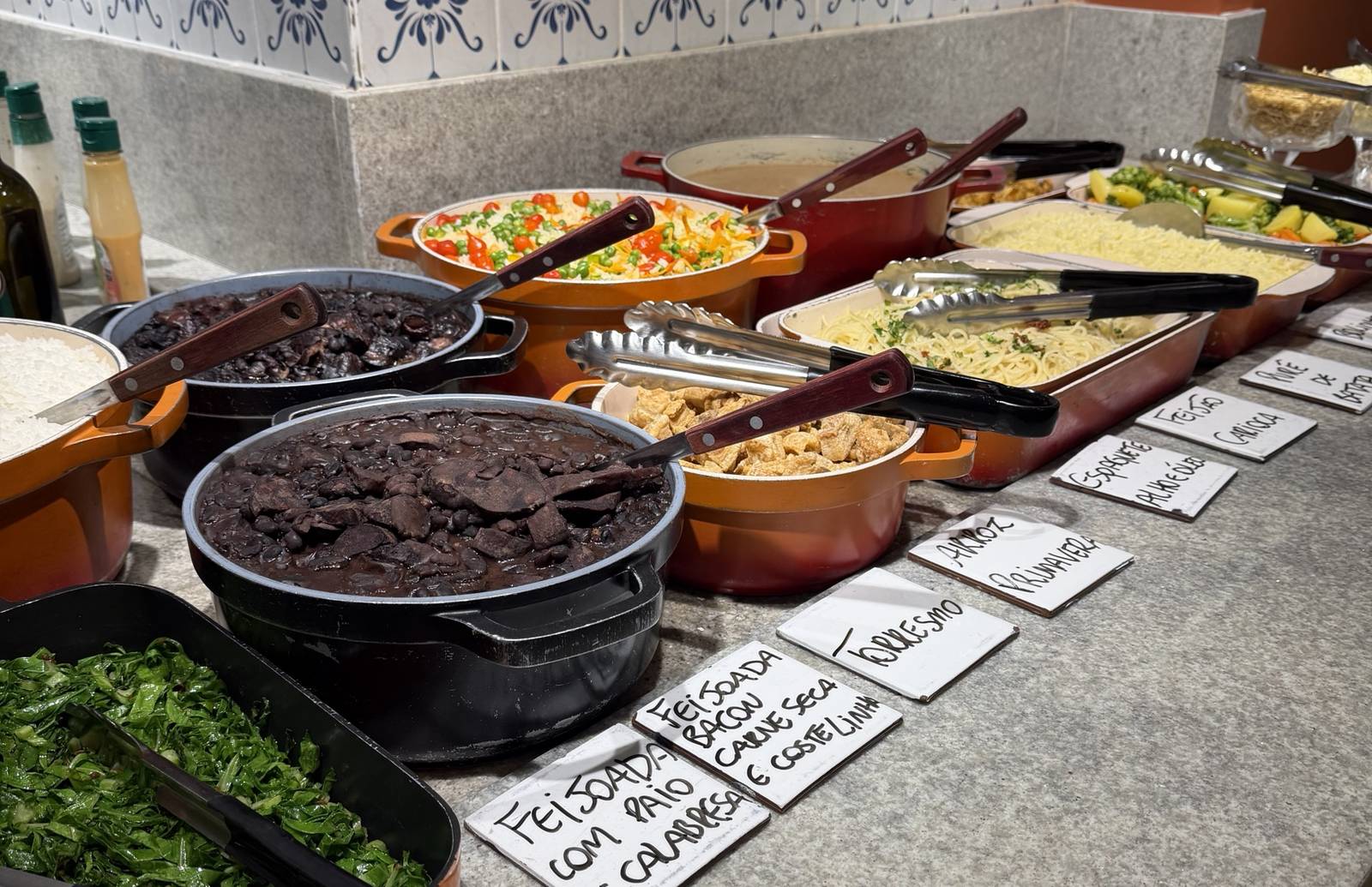 Buffet com panelas de feijoada, arroz primavera, torresmo e couve, com as plaquinhas escritas à mão